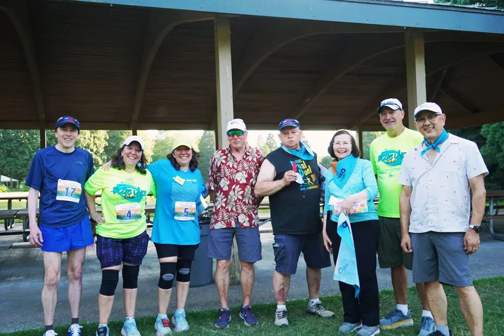 A group of eight people, some wearing race bibs and bandanas, posing for a photo under a covered pavilion at an outdoor event.