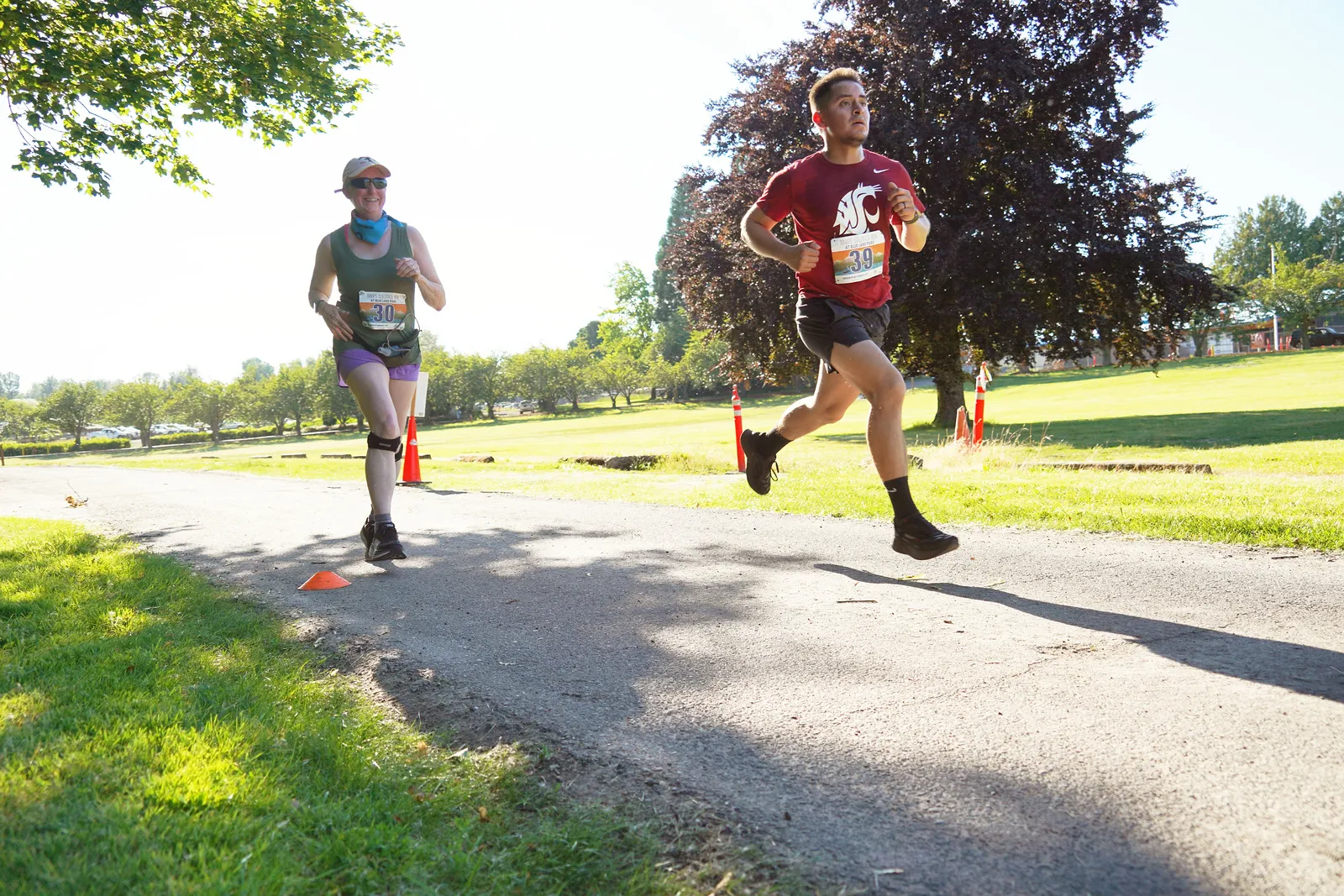 Two runners, one in a red shirt and one in a green tank top, running on a paved path in a park.