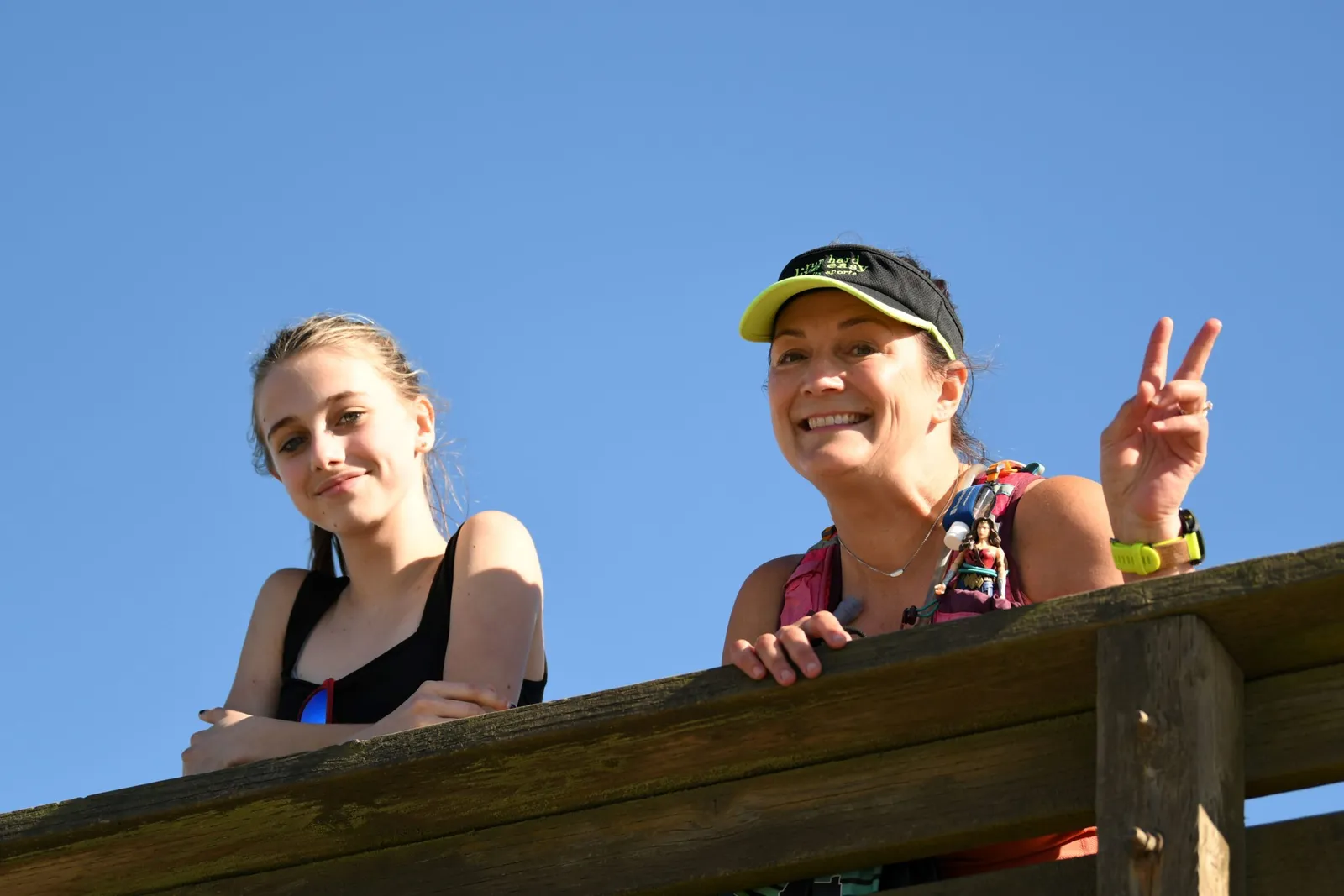 Two women smiling and looking up, one making a peace sign, against a clear blue sky.