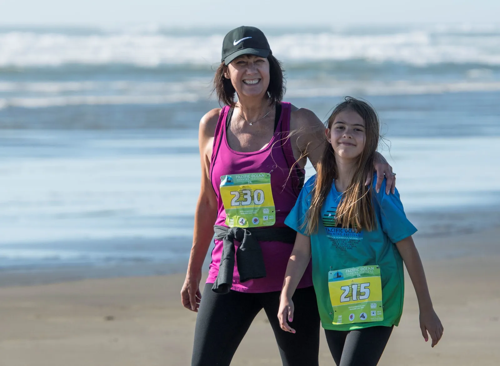 A woman and a young girl smile at the camera on a beach, both wearing race bibs.