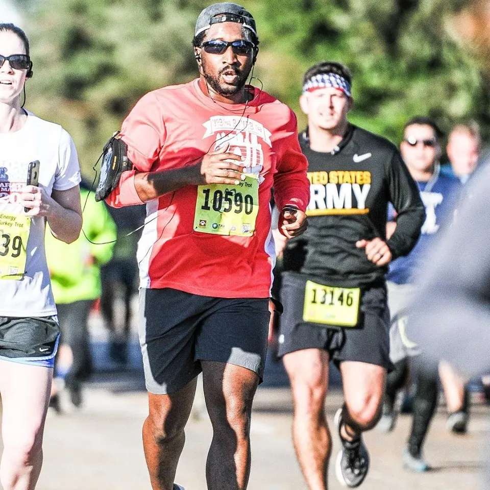 A male runner in a red long-sleeved shirt and black shorts runs in the foreground, wearing sunglasses and a baseball cap. Other runners are visible behind him.