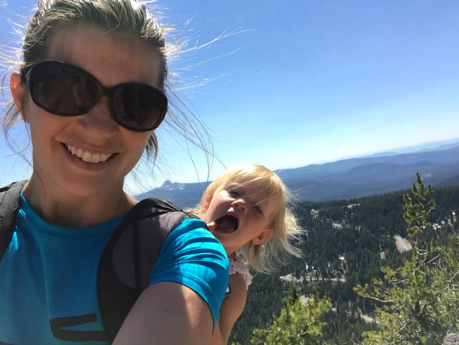 A woman smiles for a selfie while carrying a child in a backpack, with a scenic mountain landscape in the background.