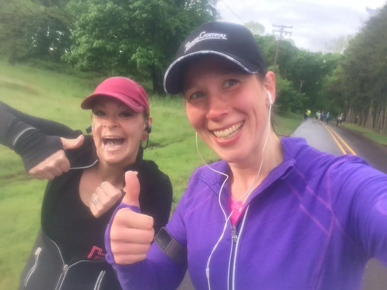Two women smiling and giving thumbs up while running on a road during a race.