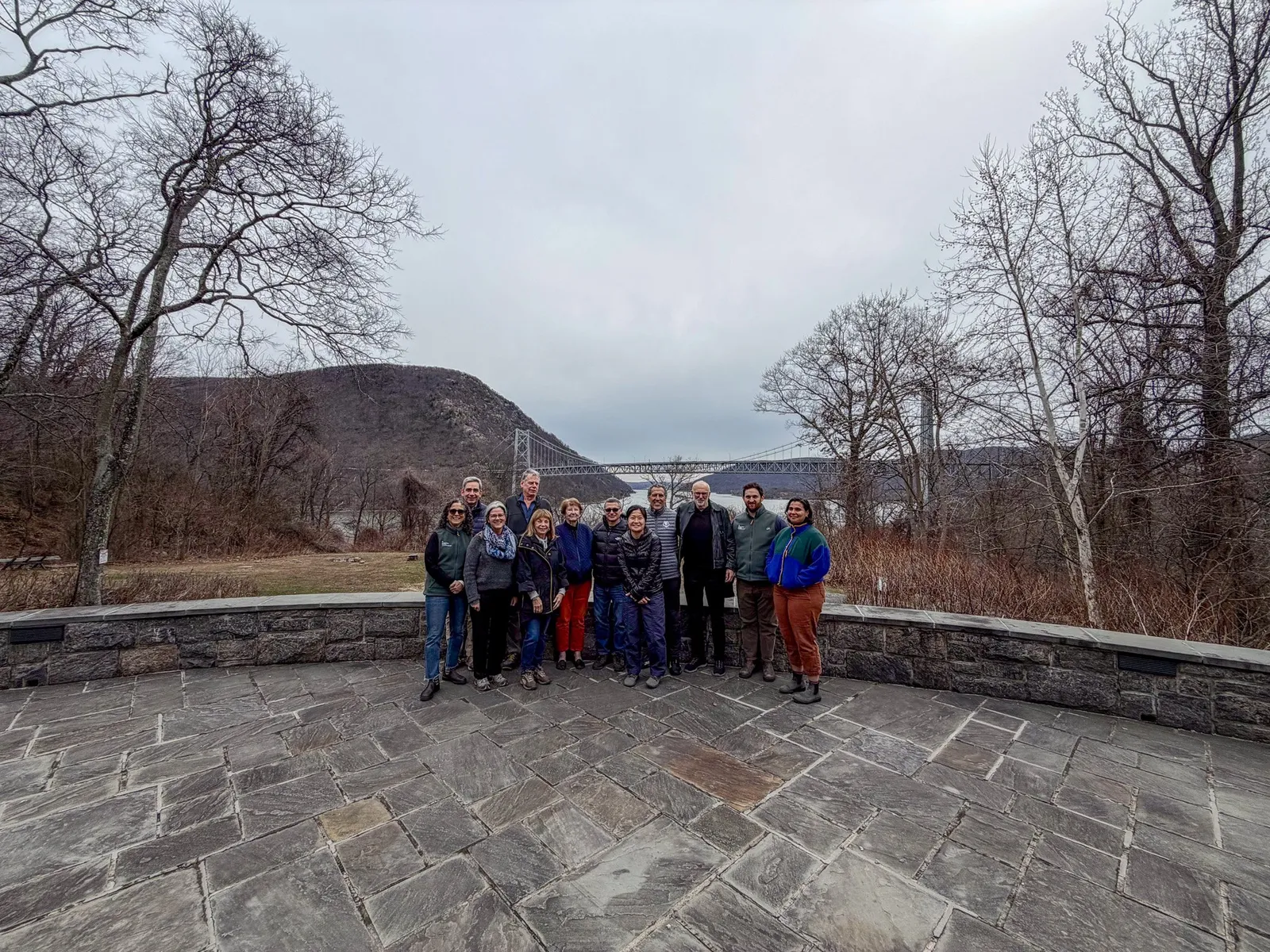 A group of people stand together on a stone patio with a bridge and river in the background.