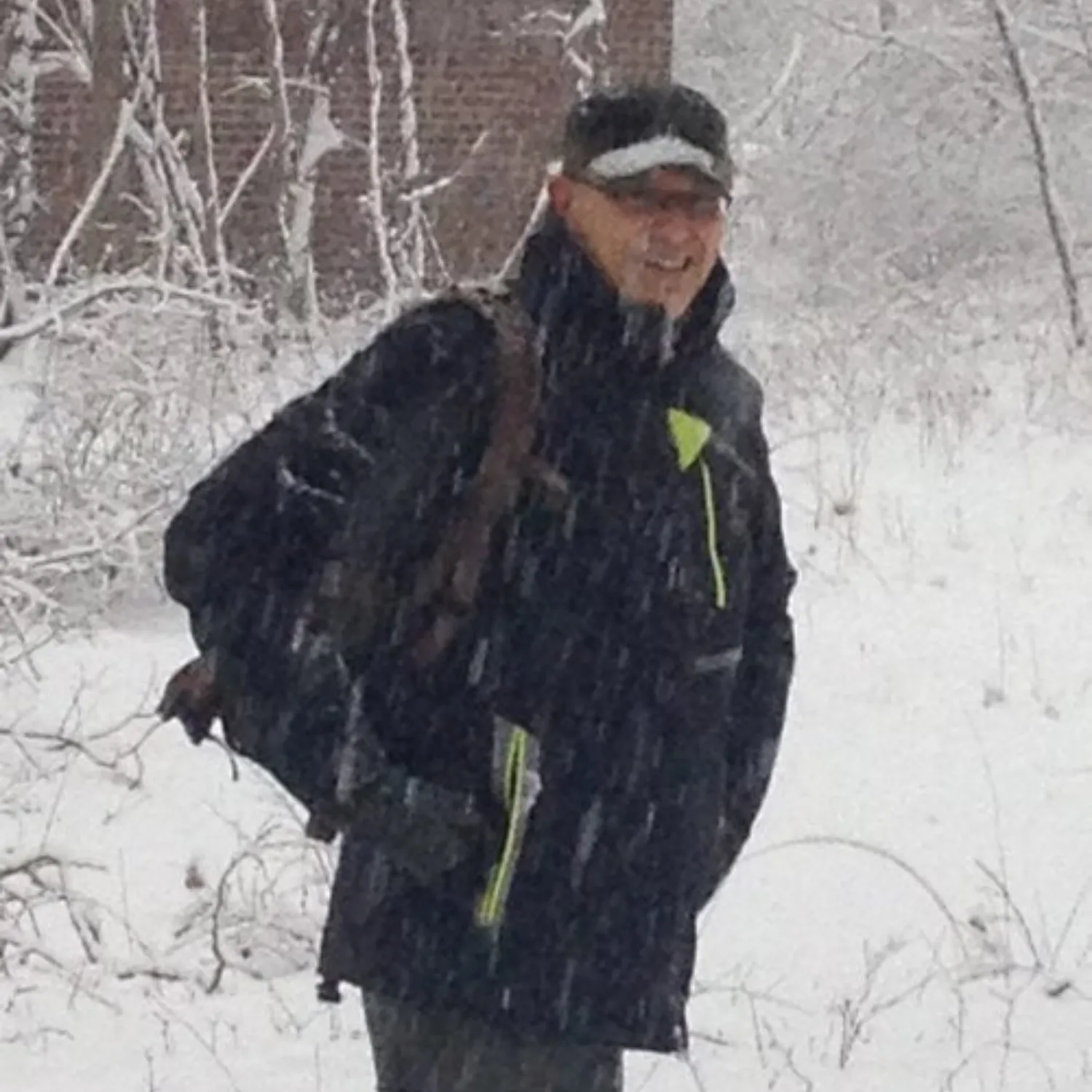 A man wearing a jacket and cap smiles while standing in a snowy forest.