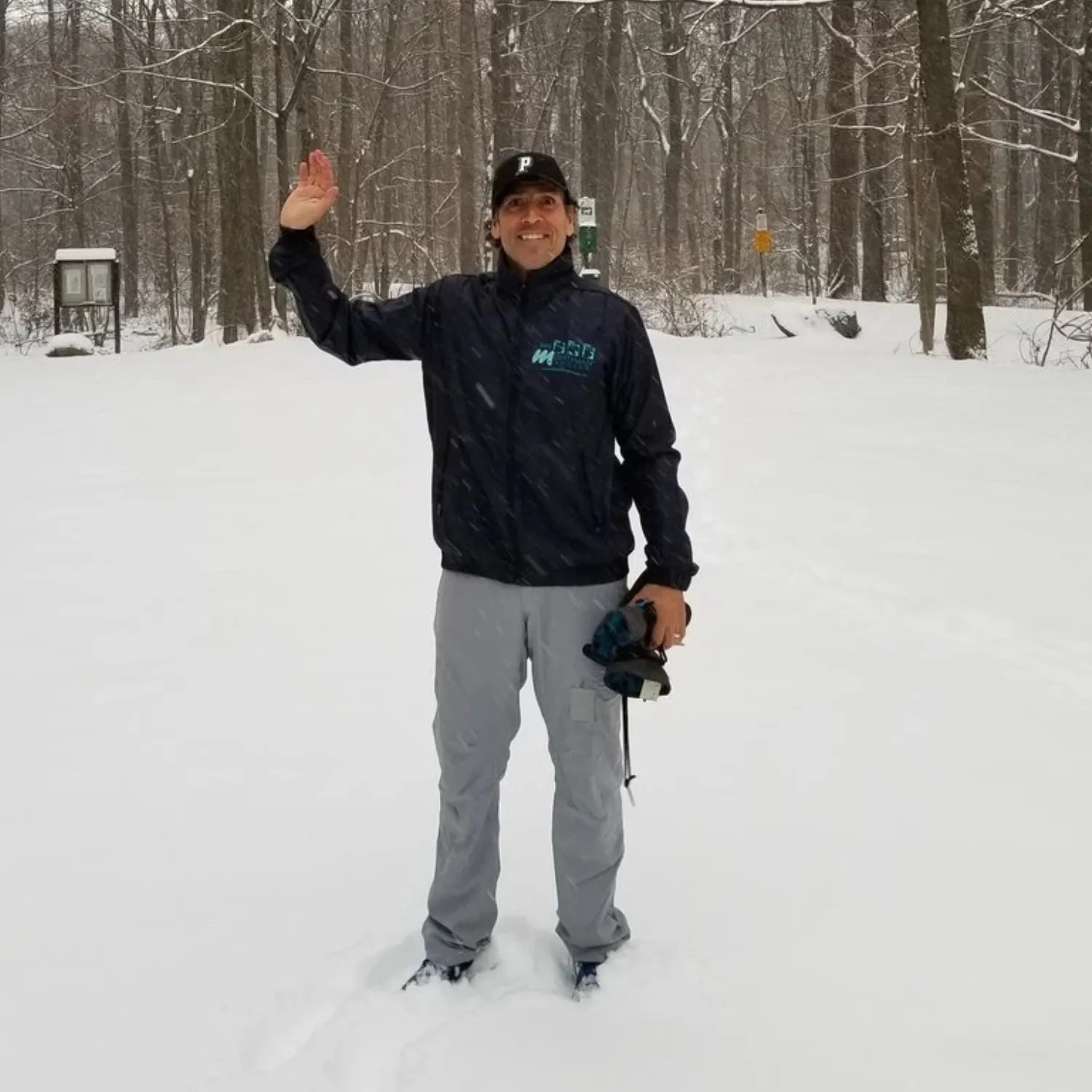 A man wearing a dark jacket and grey pants waves while standing in the snow in a wooded area.
