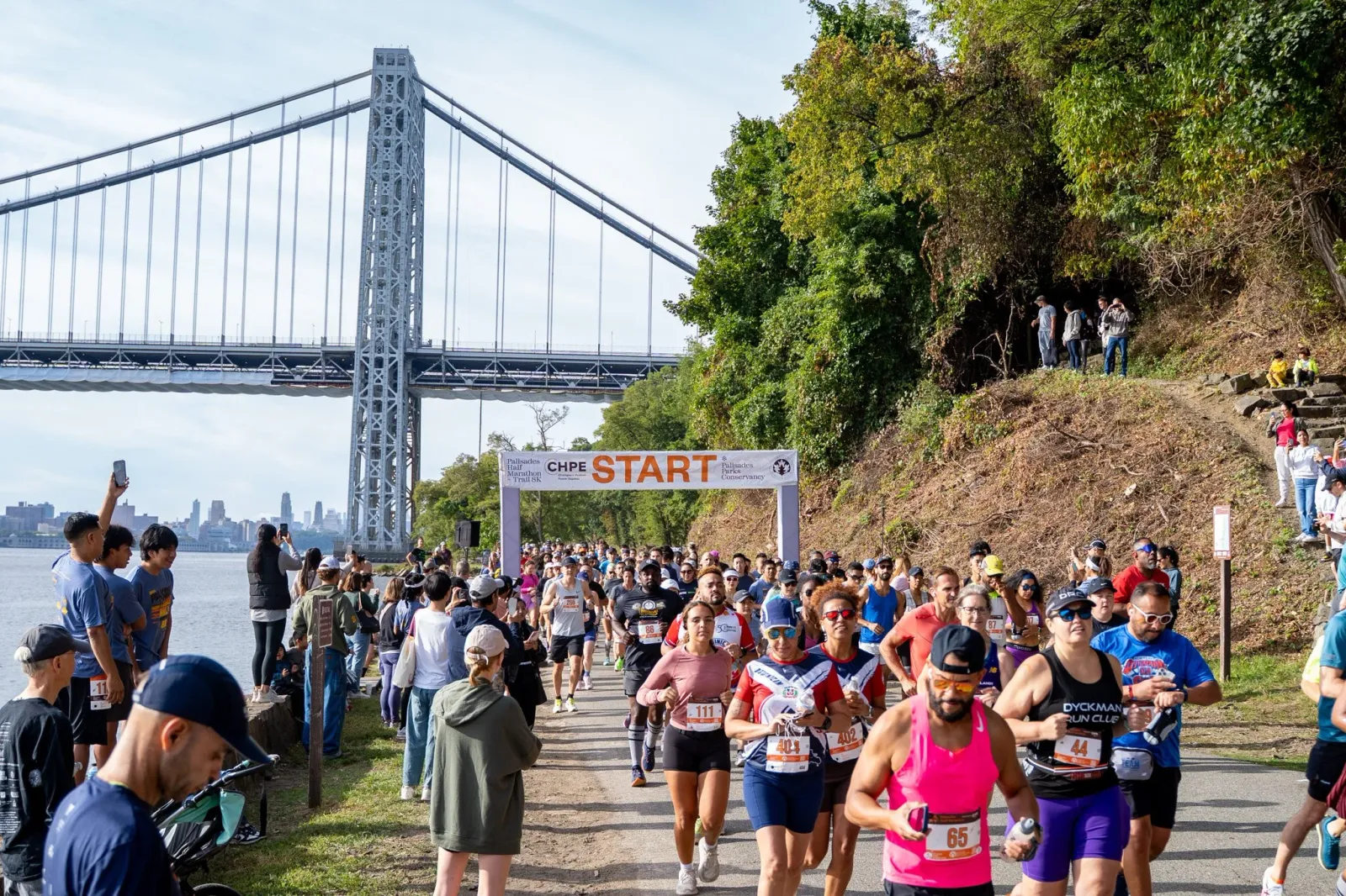 Runners starting the Palisades Half Marathon and 6K Trail Run with a large suspension bridge in the background.