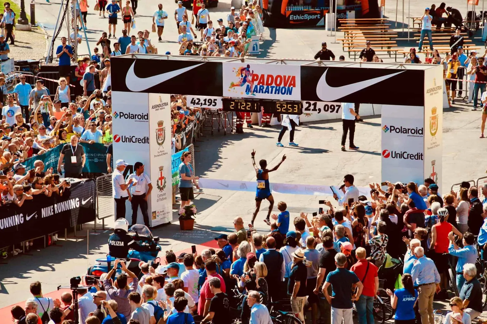 A runner crosses the finish line of the Padova Marathon with arms raised in celebration, cheered on by a large crowd.