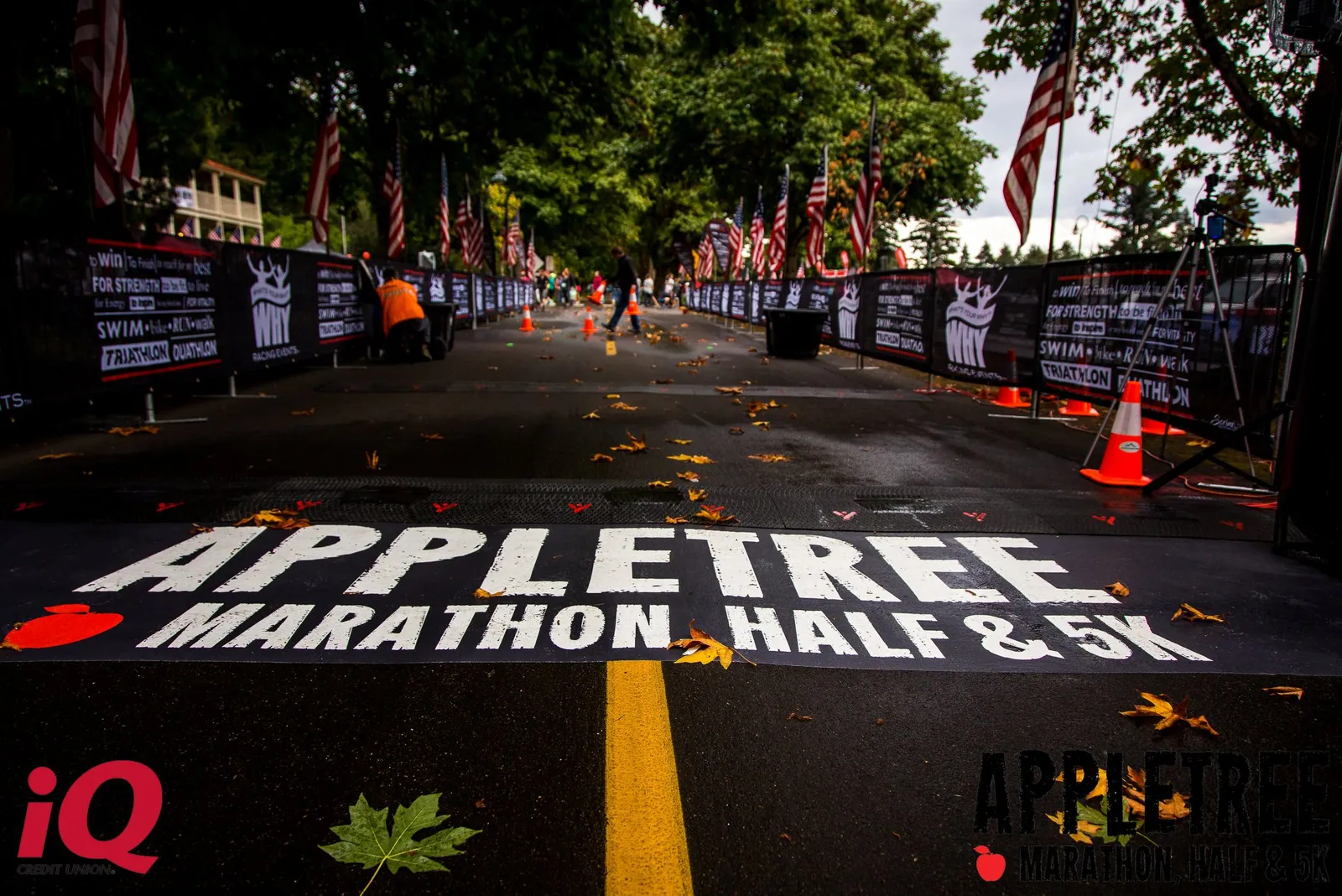 The start/finish line of the Appletree Marathon, Half & 5K, with American flags and banners lining the course, and a crowd of runners in the background.