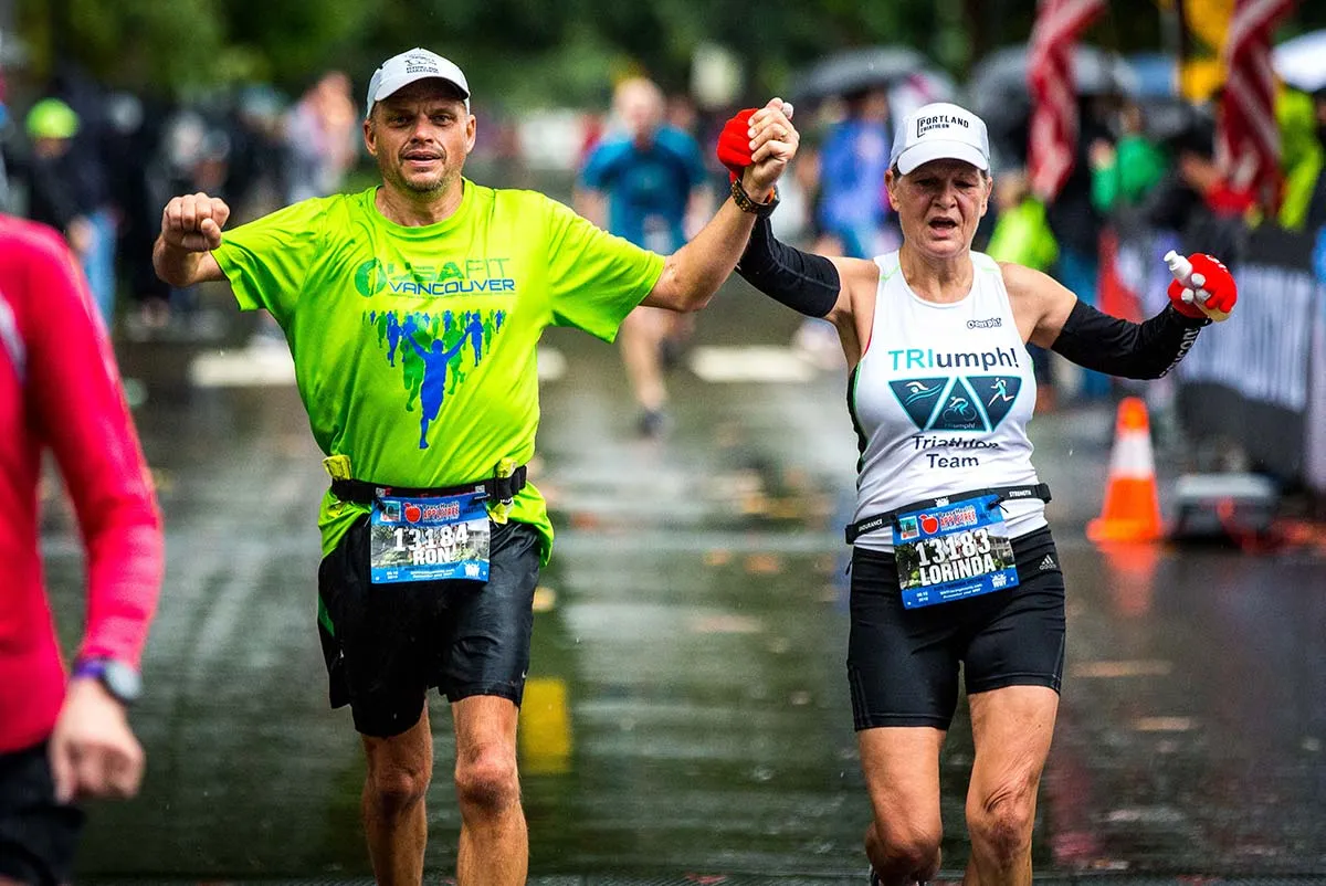 Two runners, a man and a woman, crossing the finish line with their arms raised in celebration, holding hands.