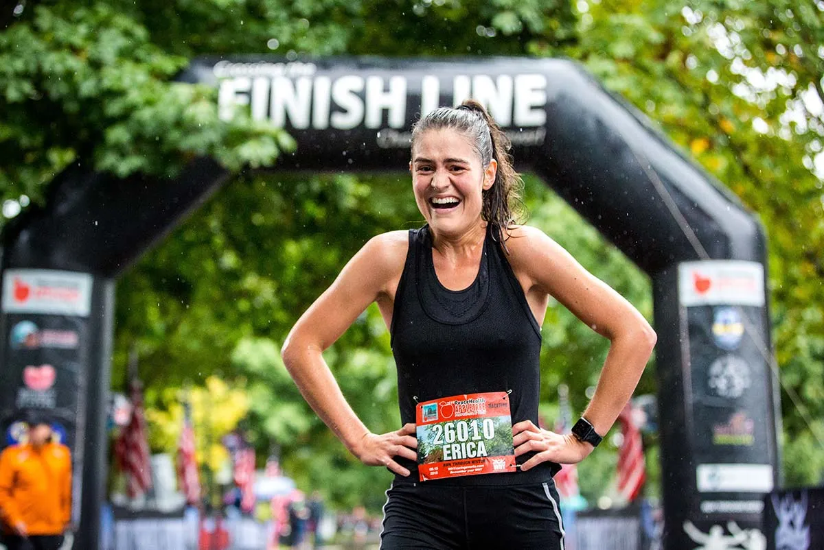 A joyful female runner with her hands on her hips crosses the finish line under an archway, with rain falling.