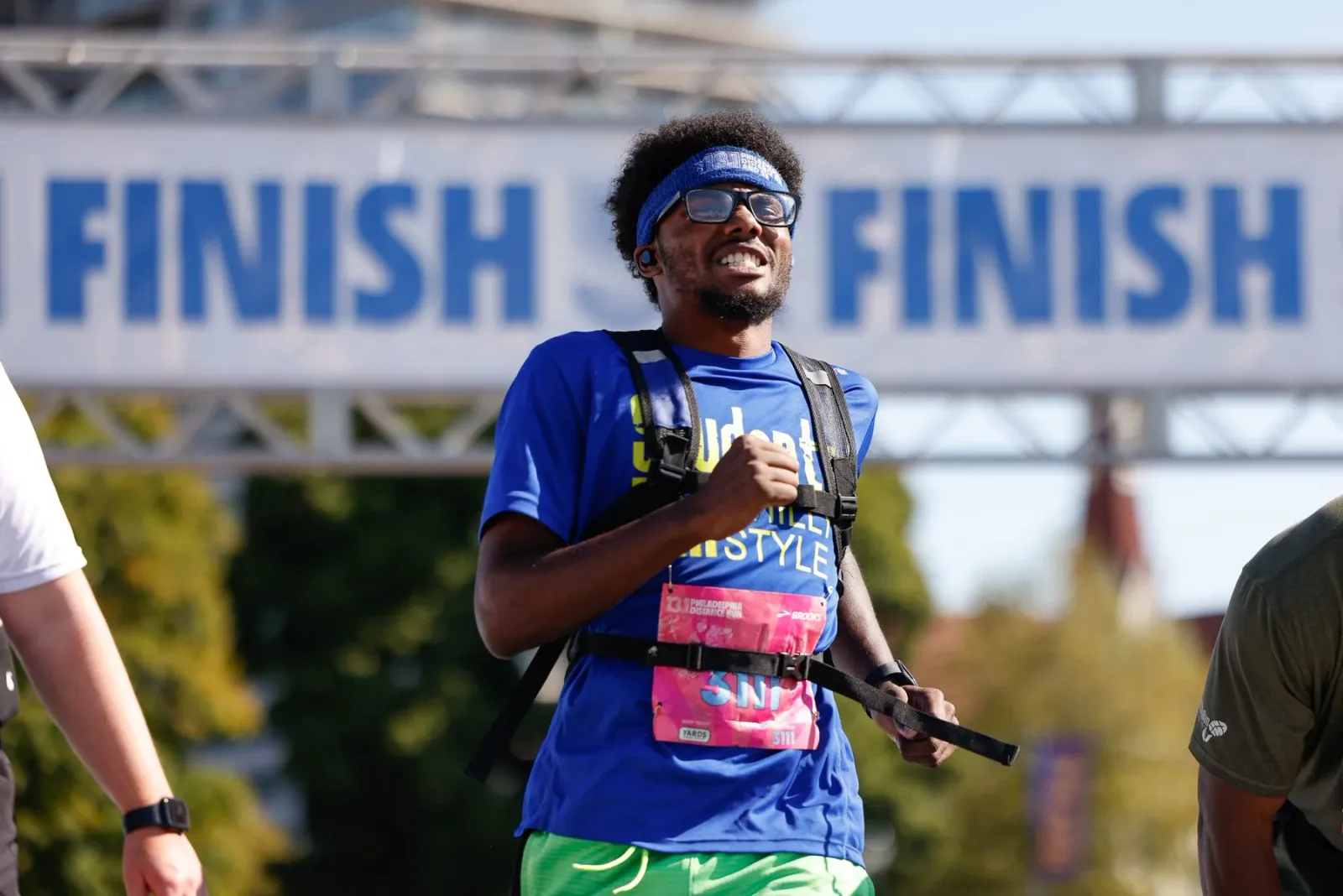 A runner with a blue shirt and green shorts runs towards the finish line, wearing glasses and a headband.