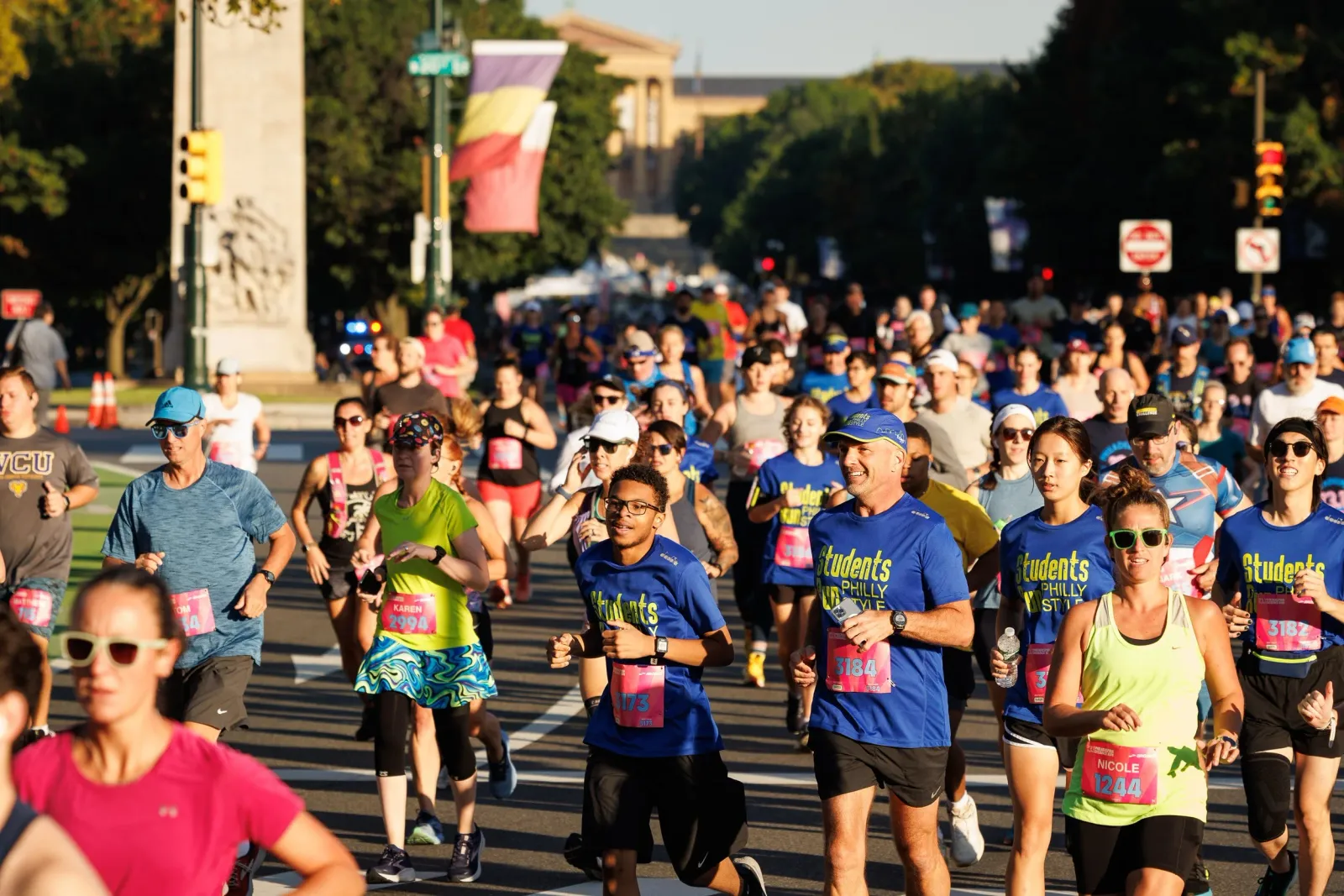 A diverse group of runners, many wearing blue t-shirts with yellow lettering, are running down a city street during a race. The sun is shining, and there are trees and buildings in the background.