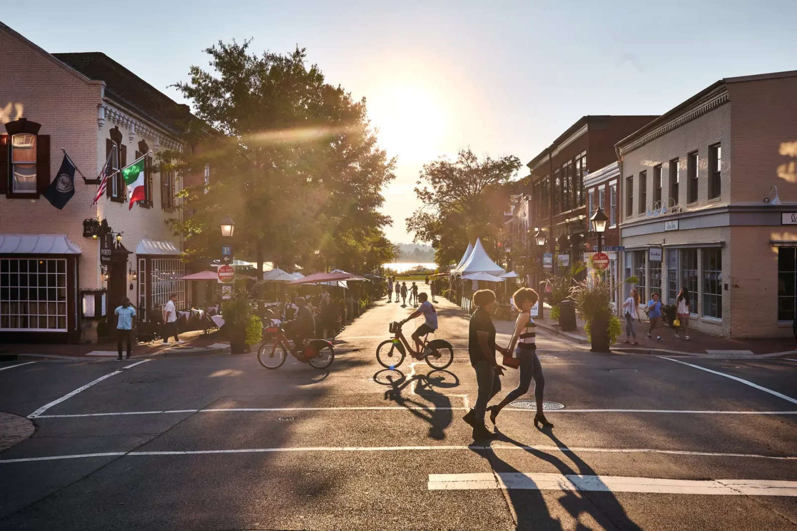 People walking and cycling on a street lined with shops and outdoor seating, with the sun setting in the background.