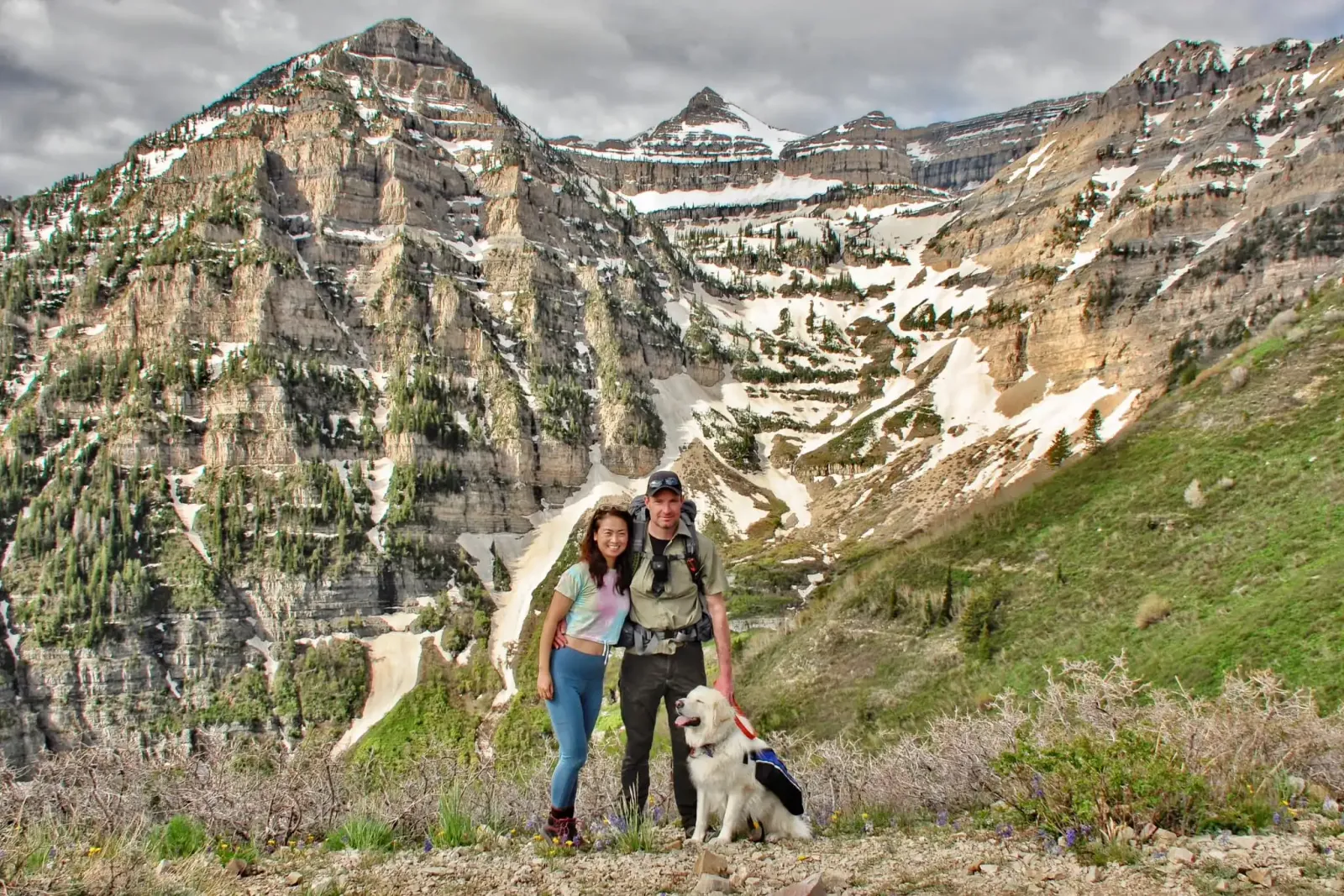 A couple and their dog pose for a photo in front of a majestic mountain range with snow-capped peaks.