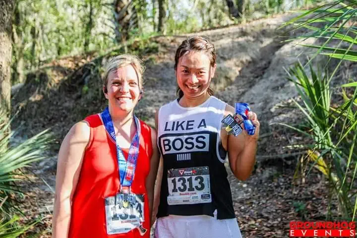 Two smiling women, one wearing a red tank top and a medal, the other wearing a 'Like a Boss' tank top and holding up medals, pose for a photo on a trail after a race.