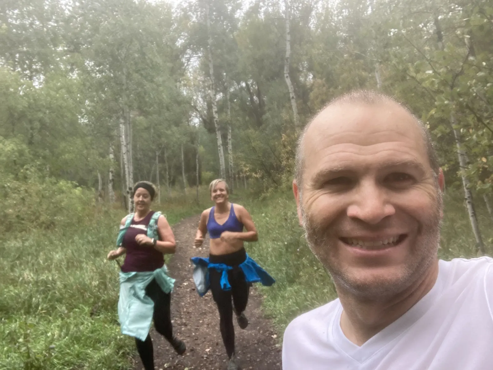 A man takes a selfie with two women running on a forest trail behind him.