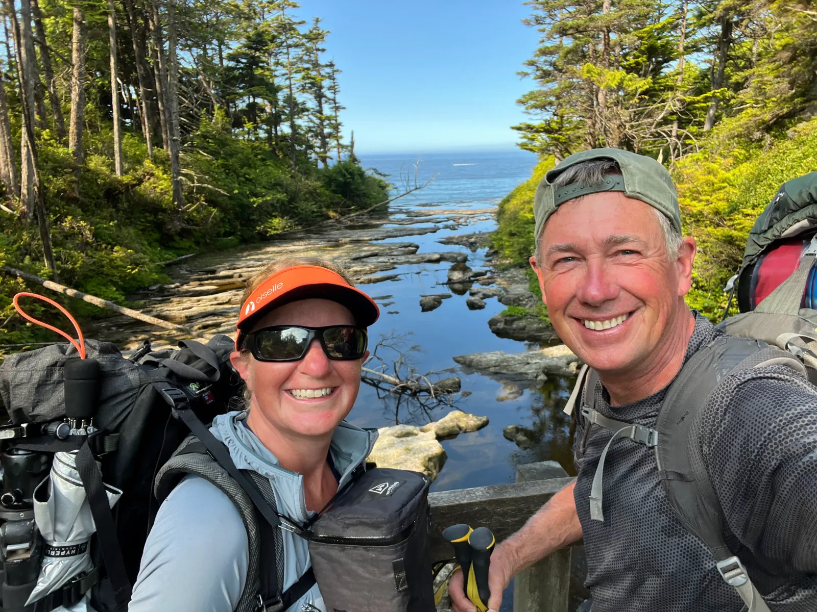 Two smiling runners, a man and a woman, wearing backpacks and athletic gear, pose for a selfie on a wooden bridge over a rocky stream with a view of the ocean and pine trees in the background.
