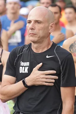A bald man with his hand over his heart stands in a crowd of runners at the start line of a race.