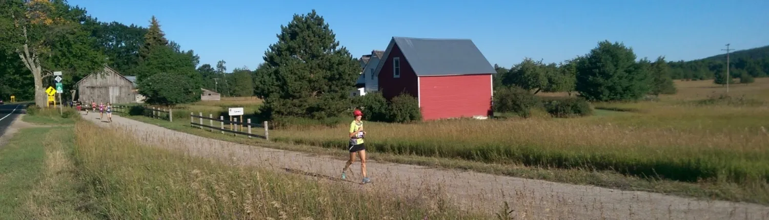 A female runner in a yellow shirt and red hat runs on a dirt path next to a grassy field, with a red barn and trees in the background.