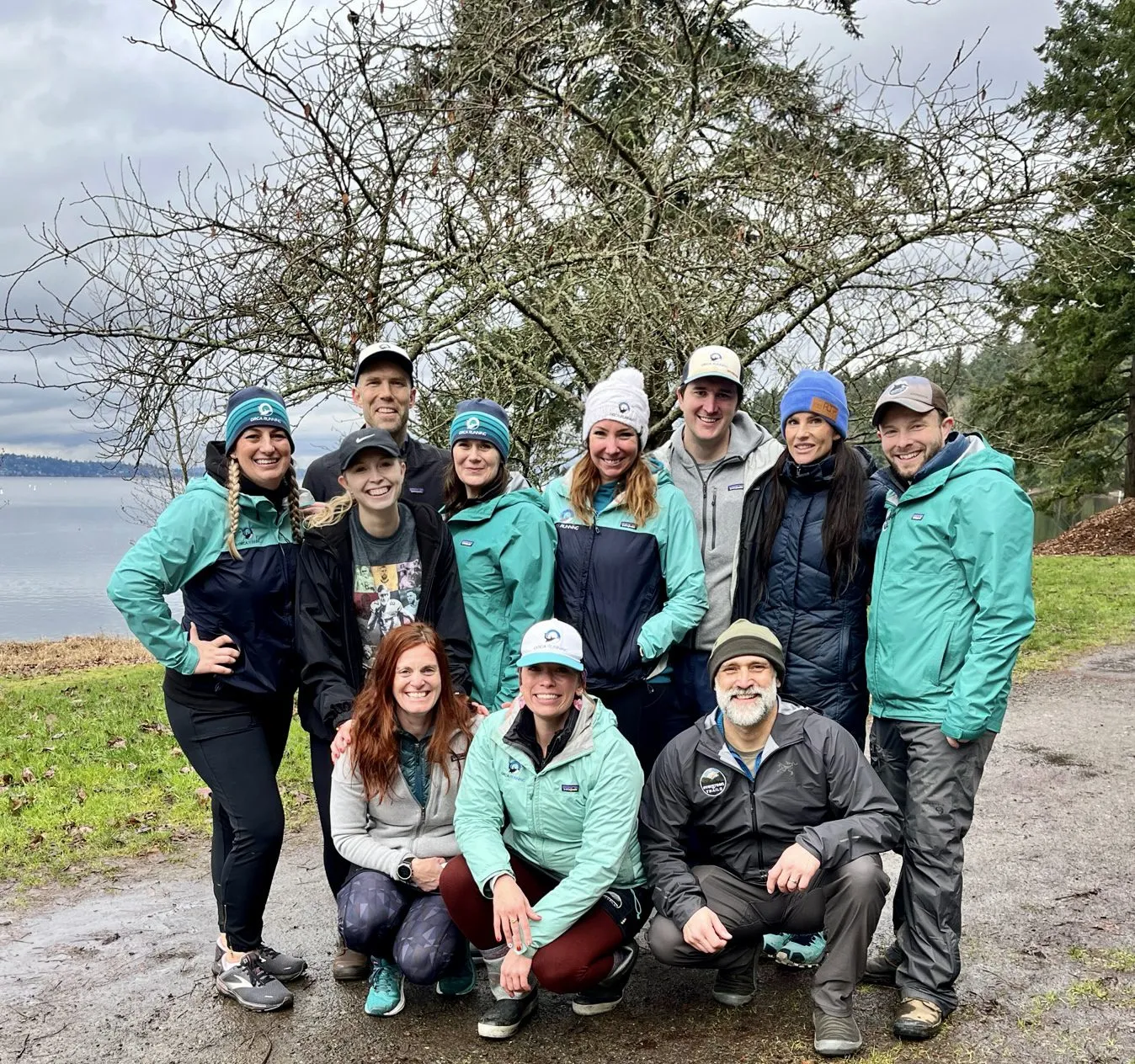 A group of ten people, some wearing matching teal jackets and beanies, pose for a photo outdoors with a body of water and trees in the background.