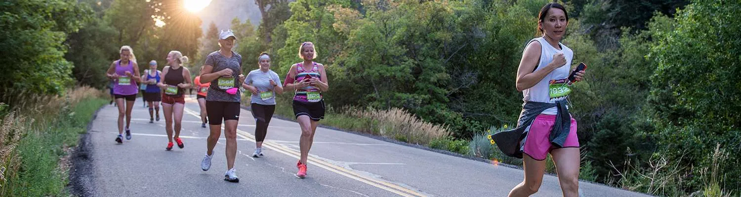 A group of women runners on a road with trees and sunlight in the background.