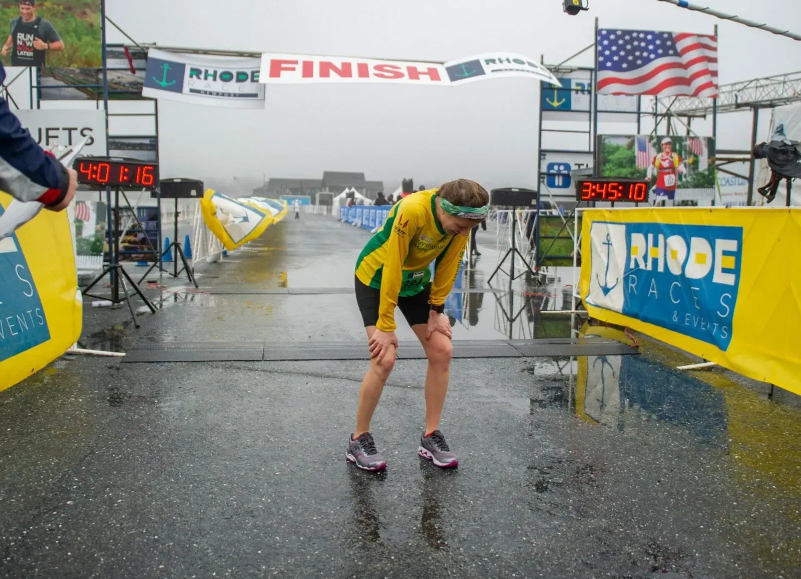 A runner bends over, hands on knees, at the finish line of a race on a wet road, with banners and clocks in the background.