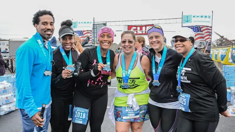 A group of six runners, all smiling and wearing medals, pose for a photo in front of a finish line banner.