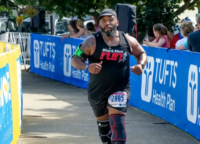 A male runner wearing a black 'Black Men Run' tank top and a race bib number 2885 runs towards the camera, with spectators and sponsor banners in the background.