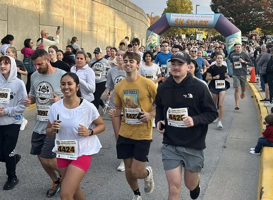 A large group of runners are starting a race under a purple and white archway that says "Purdue Boilermaker Half Marathon 5K Start".