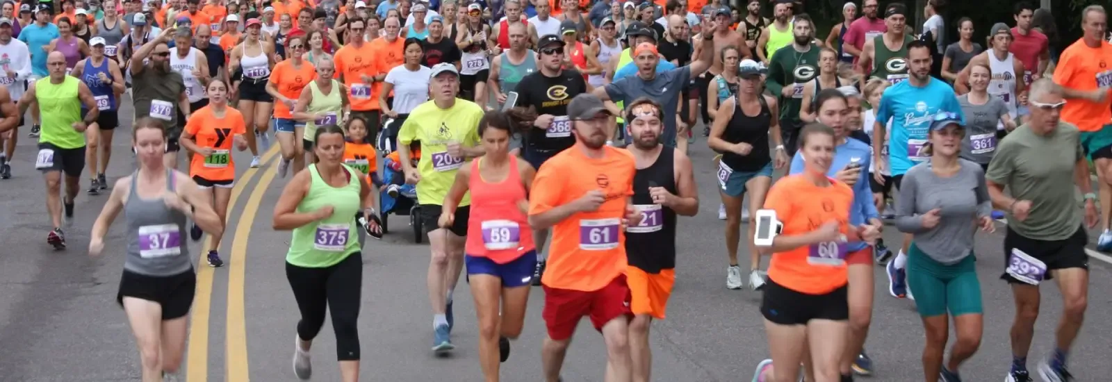 A large group of runners of various ages and genders are running down a road during a race, many wearing bright colored shirts.