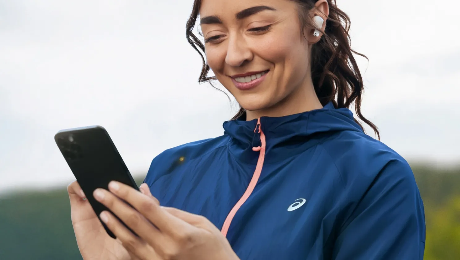 A woman with earbuds smiles as she looks at her smartphone while wearing a blue running jacket.