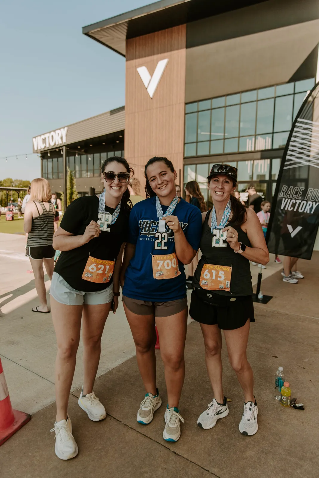 Three smiling women wearing race medals and bibs stand together outside a building with a large 'VICTORY' sign.