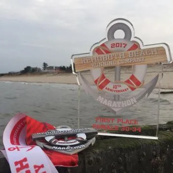 Rehoboth Beach Running Company 10th Anniversary Marathon first place trophy and medals displayed on a wooden post with a beach and ocean in the background.