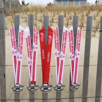 Five race medals with red and white ribbons hanging on a wooden fence, with a beach and dunes in the background.