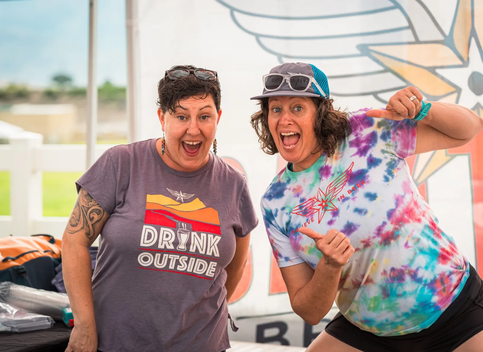 Two women with excited expressions, one wearing a grey t-shirt with 'DRINK OUTSIDE' and the other a tie-dye t-shirt, posing for a photo at an outdoor event.