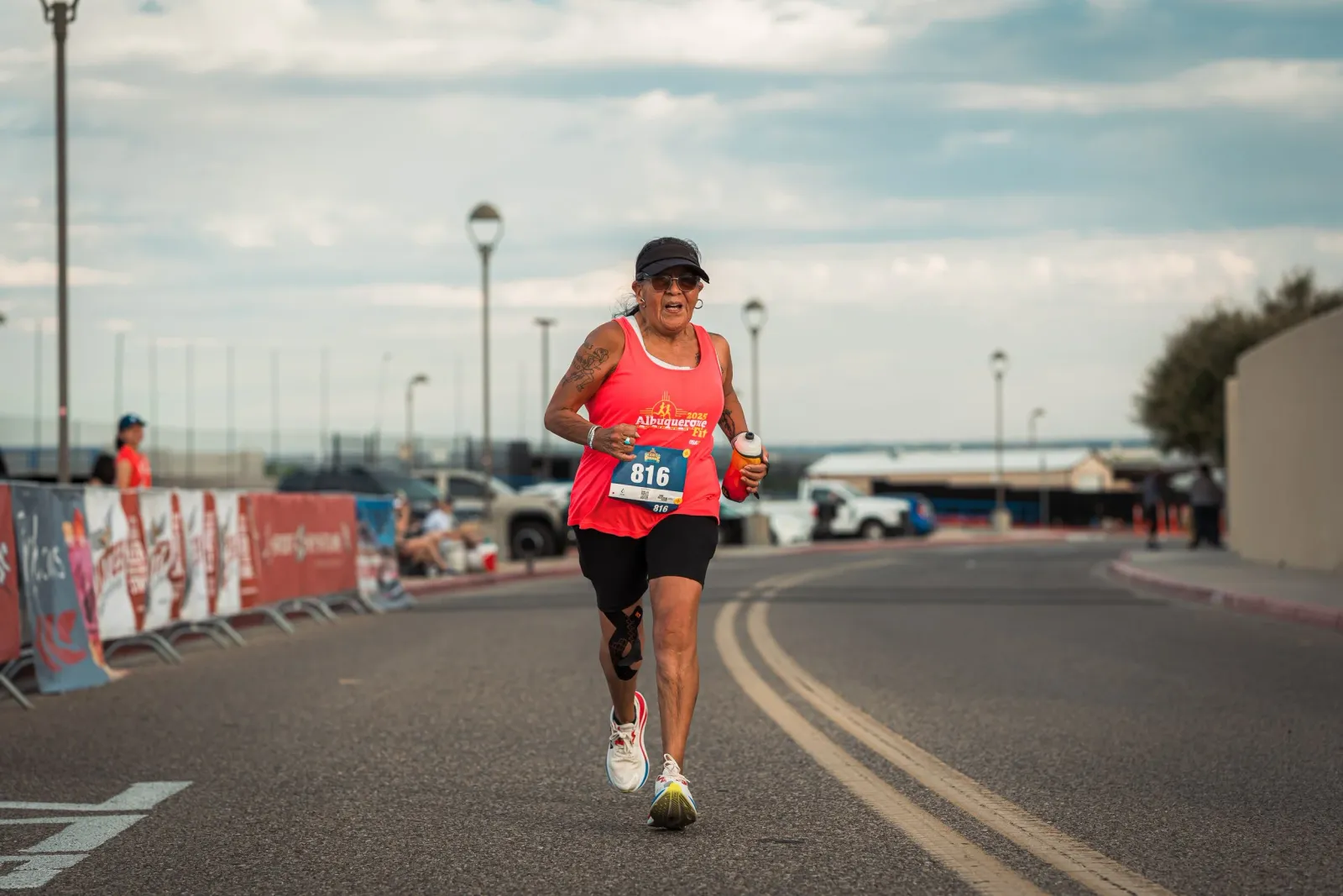 A female runner in a bright pink tank top and black shorts runs on a paved road, wearing a bib with the number 816 and a black baseball cap.