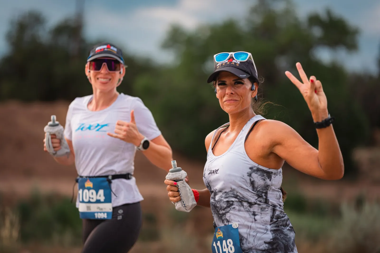Two female runners smiling and giving thumbs up and peace signs while running a race.