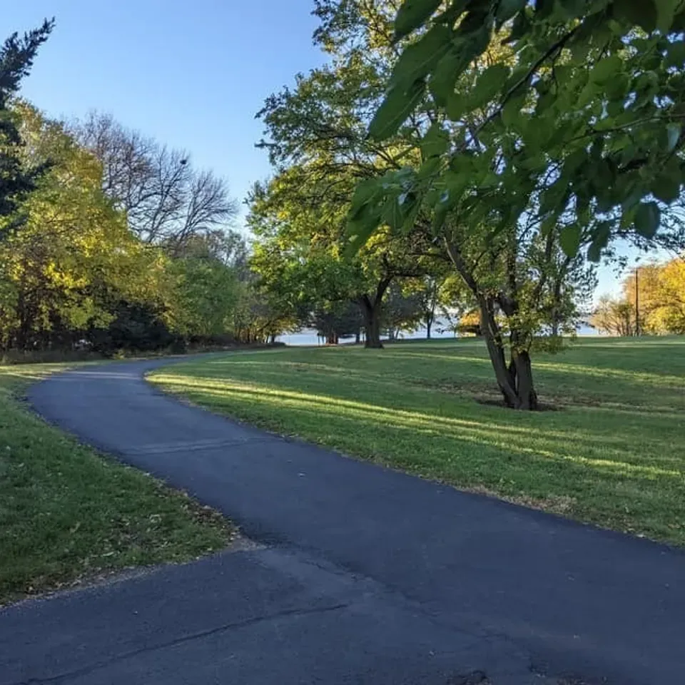 A paved path curves through a grassy park with trees and a body of water in the distance under a clear blue sky.