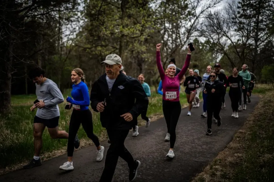 A group of runners on a paved path through a wooded area, with one runner in the foreground raising her arms in celebration.
