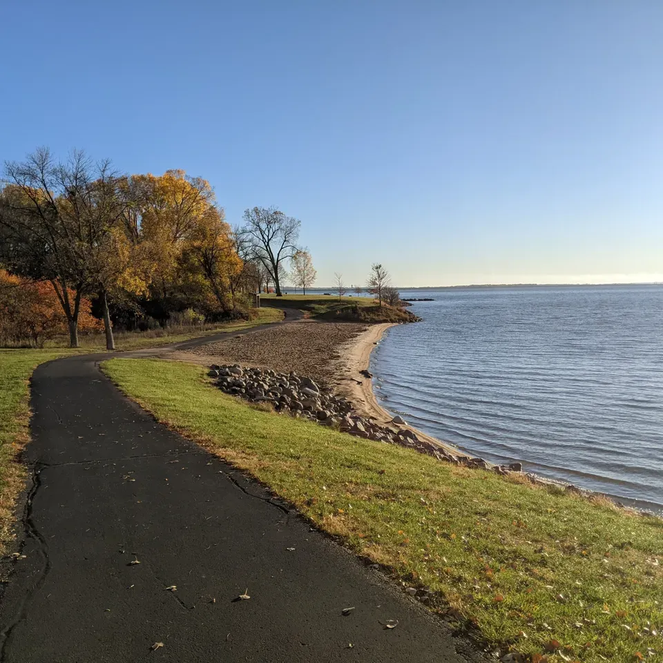 A paved path curves along a grassy hill next to a sandy beach and a large body of water under a clear blue sky.