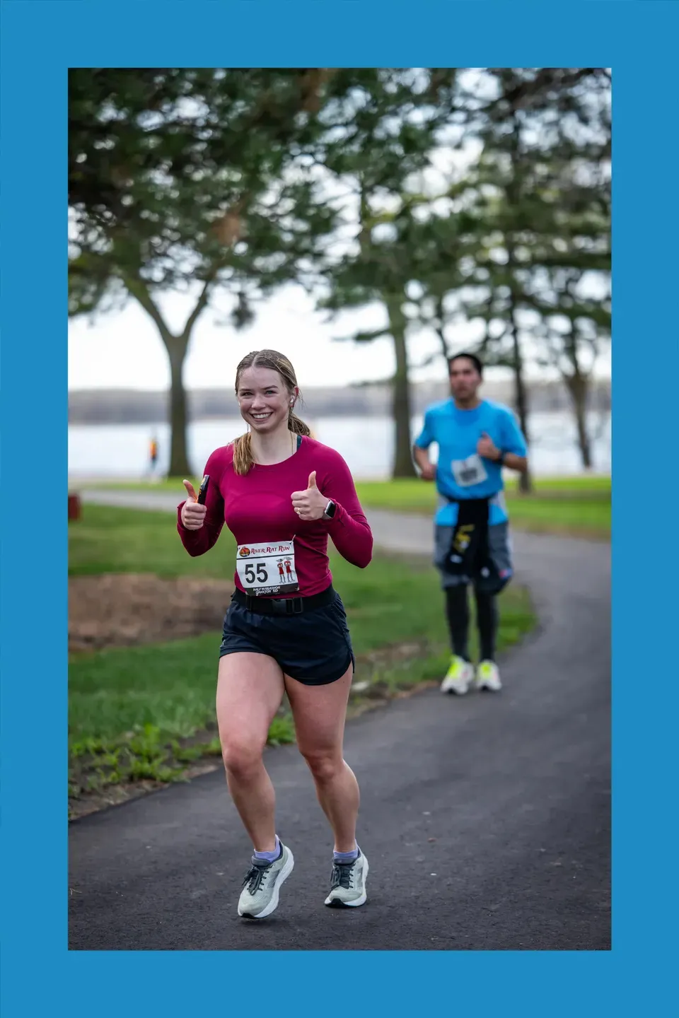 A female runner smiles and gives two thumbs up while running on a paved path, with another runner in the background.