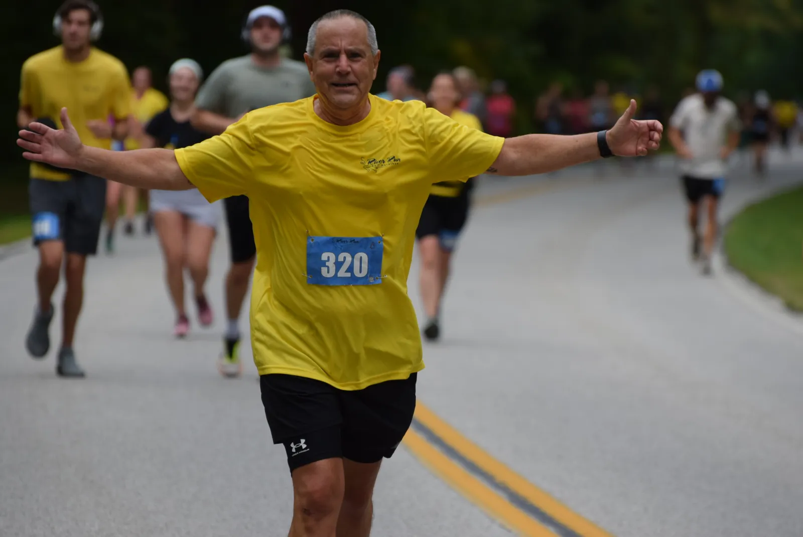 A male runner in a yellow shirt with bib number 320 and arms outstretched, running on a road with other runners in the background.