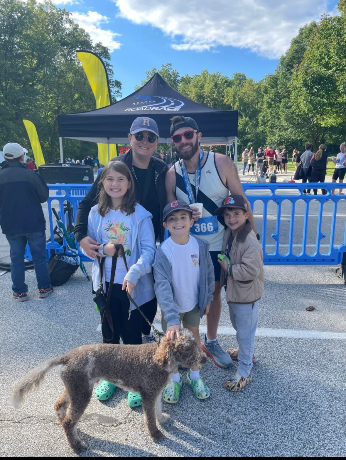 A family with a dog poses for a photo in front of a race tent, with other runners in the background.