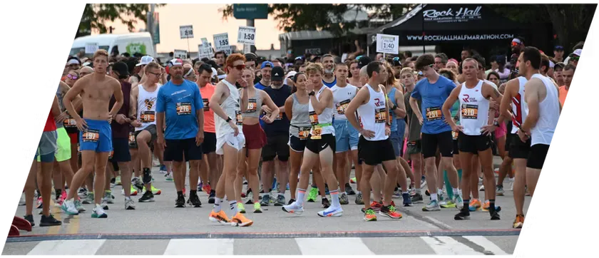 A diverse group of runners in athletic wear stand at the start line of a race, with bib numbers visible, ready to begin.
