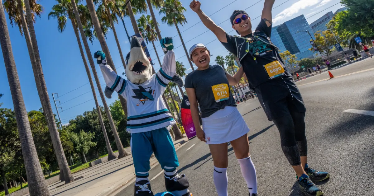 A mascot and two runners celebrating with their arms in the air during a road race, with palm trees and city buildings in the background.