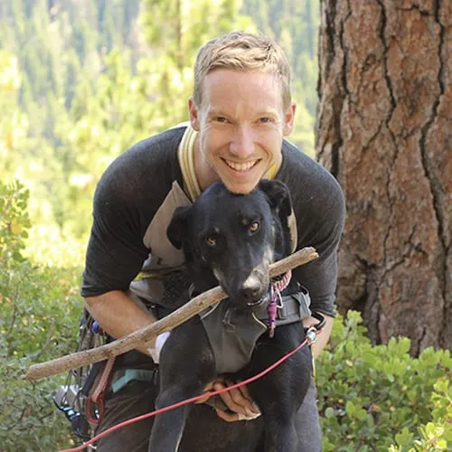 A smiling man holding a black dog with a stick in its mouth, with a forest background.