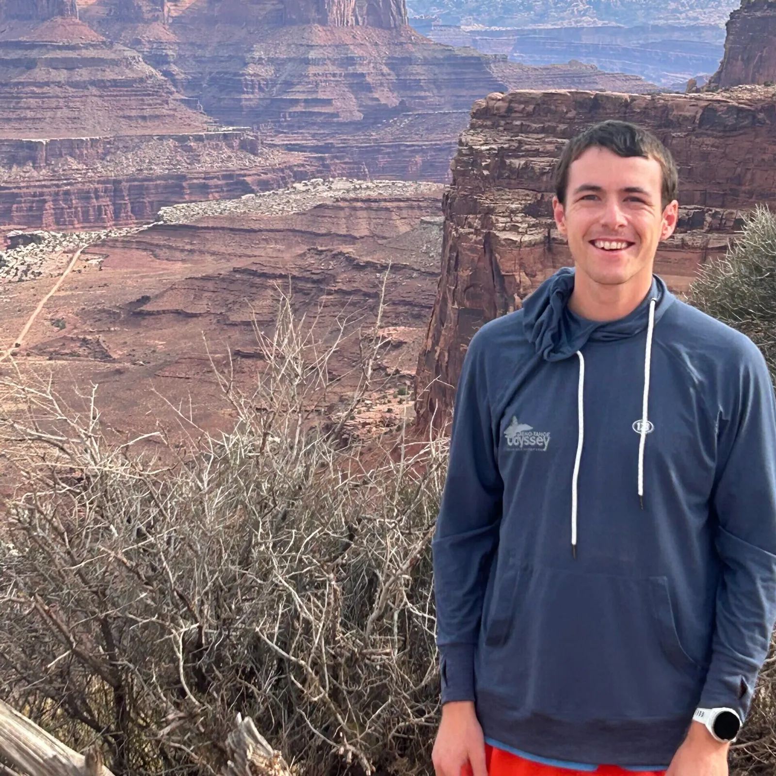 A smiling man in a blue hoodie stands in front of a canyon landscape.