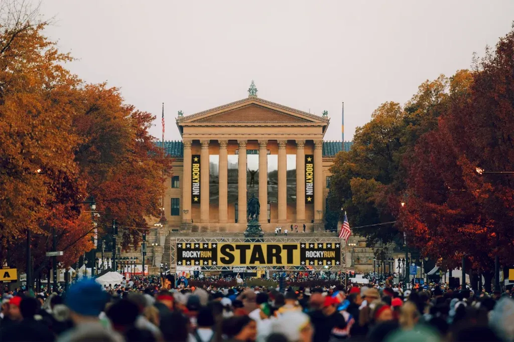 A large crowd of runners gathered at the start line in front of the Philadelphia Museum of Art, with banners reading 'ROCKY RUN' and 'START'.