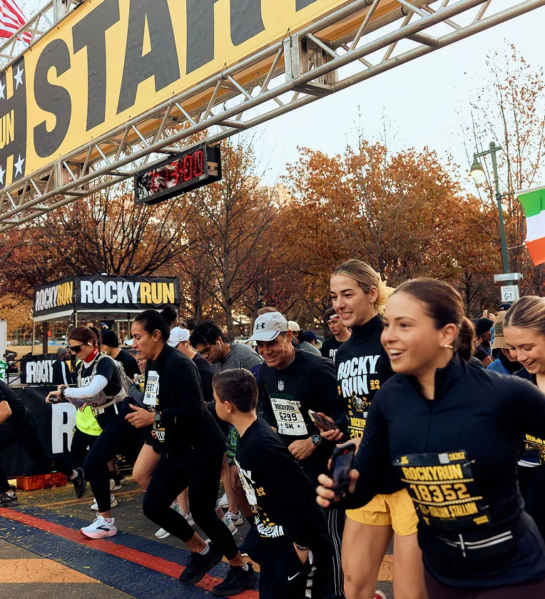 Runners at the start line of the Rocky Run race, with a large 'START' banner overhead and a digital timer displaying '0:00'.